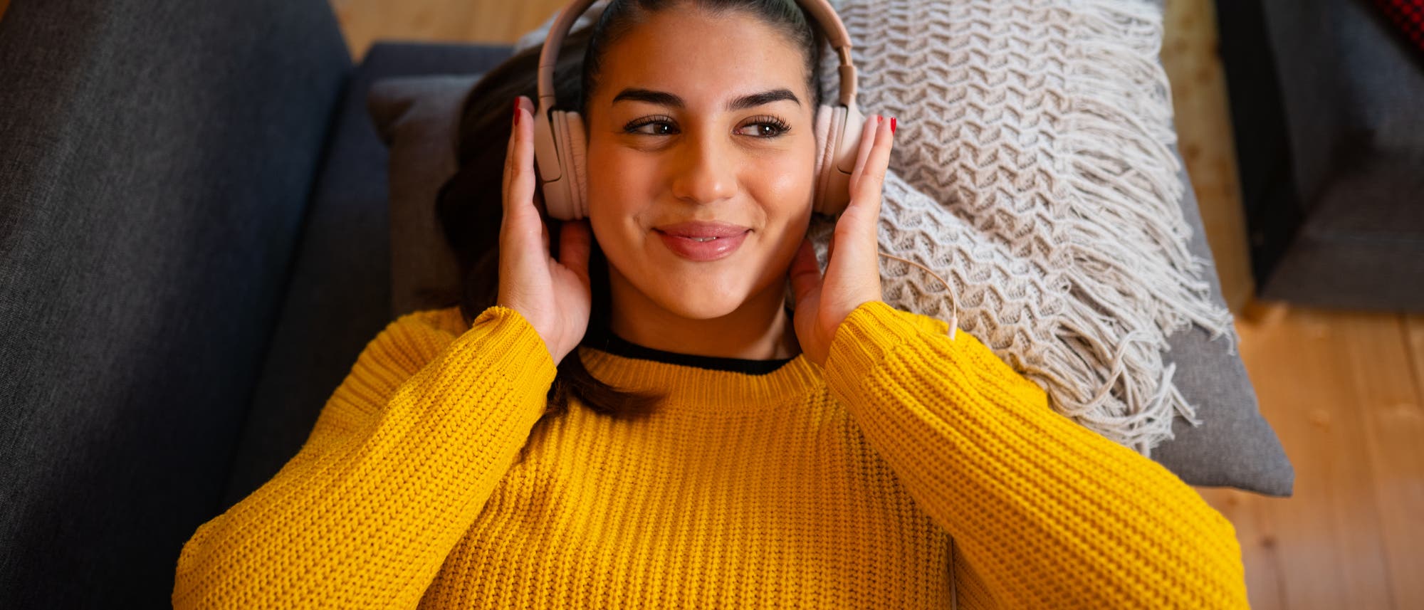 Eine Person liegt entspannt auf einem Sofa und hört Musik mit großen Kopfhörern. Sie trägt einen gelben Pullover und lächelt leicht. Neben ihr liegt ein dekoratives Kissen mit Fransen. Der Raum hat einen Holzboden und wirkt gemütlich. Eine Person liegt entspannt auf einem Sofa und hört Musik mit großen Kopfhörern. Sie trägt einen gelben Pullover und lächelt leicht. Neben ihr liegt ein dekoratives Kissen mit Fransen. Der Raum hat einen Holzboden und wirkt gemütlich.