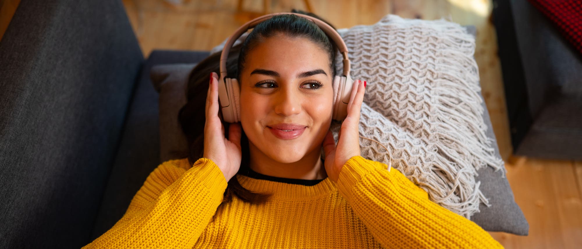Eine Frau liegt entspannt auf einem Sofa und hört Musik mit großen Kopfhörern. Sie trägt einen gelben Pullover und lächelt leicht. Neben ihr liegt ein dekoratives Kissen mit Fransen. Der Raum hat einen Holzboden und wirkt gemütlich.