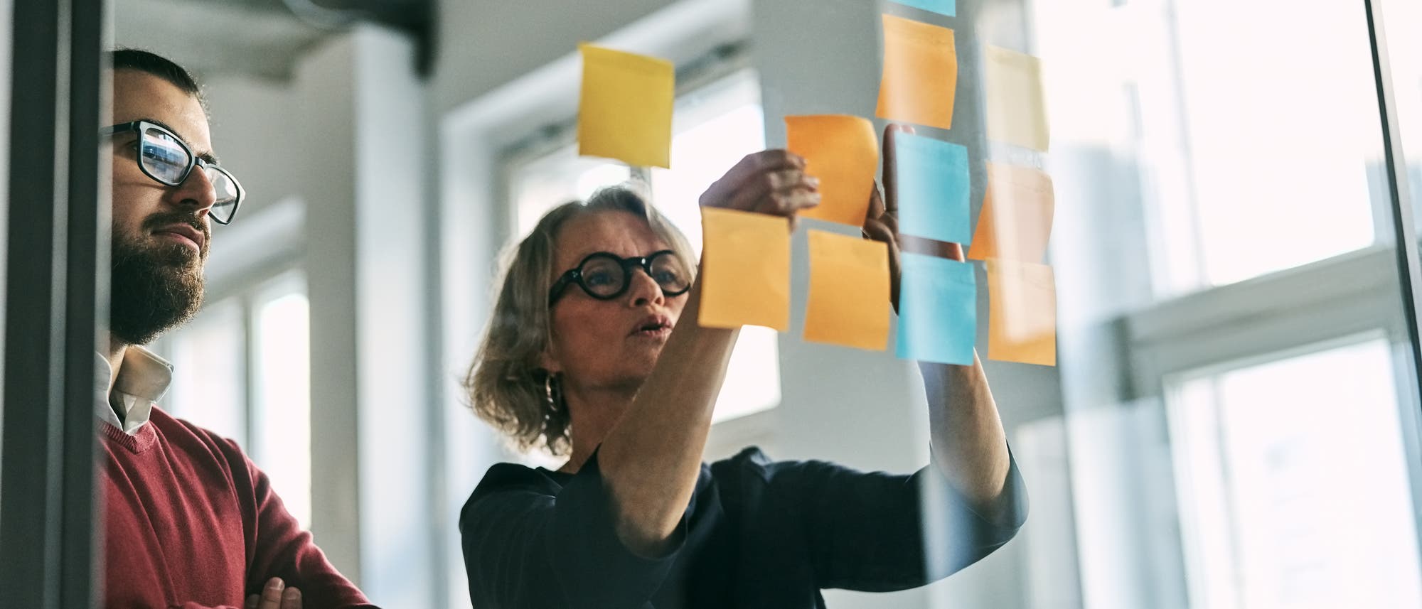 Zwei Personen stehen vor einer Glaswand, die mit bunten Haftnotizen bedeckt ist. Eine Person zeigt auf eine der Notizen, w&auml;hrend die andere aufmerksam zusieht. Die Szene findet in einem hellen Raum mit gro&szlig;en Fenstern statt. Die Notizen sind in verschiedenen Farben wie Gelb, Orange und Blau gehalten.