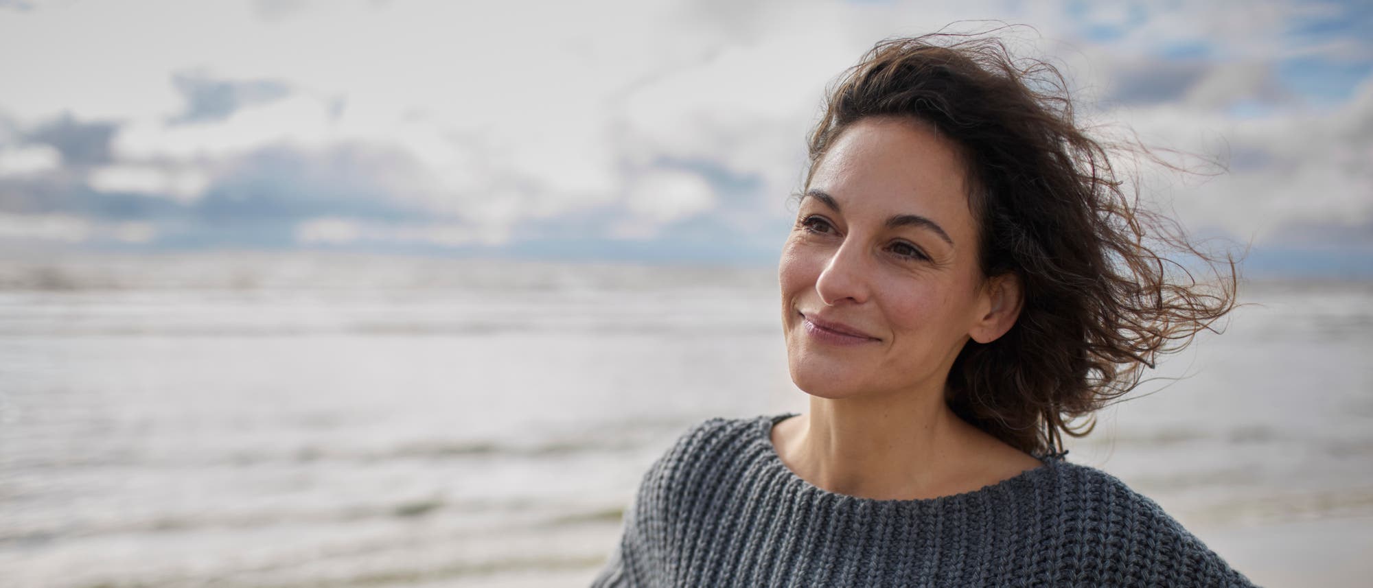 Eine Person steht am Strand und blickt l&auml;chelnd in die Ferne. Der Himmel ist bew&ouml;lkt, und das Meer ist im Hintergrund zu sehen. Die Person tr&auml;gt einen grauen Strickpullover, und der Wind weht durch ihr Haar.