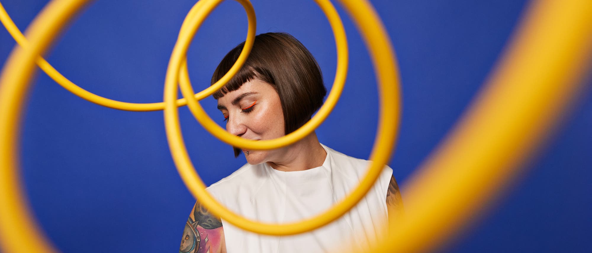 Eine Person mit einem weißen Oberteil steht vor einem blauen Hintergrund. Gelbe Spiralen verlaufen im Vordergrund des Bildes und erzeugen einen dynamischen visuellen Effekt. Die Person hat einen tätowierten Arm und schaut nach unten, was eine nachdenkliche Stimmung vermittelt. Eine Person mit einem weißen Oberteil steht vor einem blauen Hintergrund. Gelbe Spiralen verlaufen im Vordergrund des Bildes und erzeugen einen dynamischen visuellen Effekt. Die Person hat einen tätowierten Arm und schaut nach unten, was eine nachdenkliche Stimmung vermittelt.