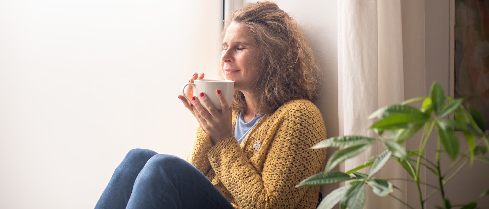 Eine Person sitzt entspannt an einem Fenster und hält eine Tasse in beiden Händen. Sie trägt einen gelben Strickpullover und hat die Augen geschlossen, während sie den Moment genießt. Im Vordergrund ist eine grüne Pflanze zu sehen. Die Szene vermittelt Ruhe und Zufriedenheit. Eine Person sitzt entspannt an einem Fenster und hält eine Tasse in beiden Händen. Sie trägt einen gelben Strickpullover und hat die Augen geschlossen, während sie den Moment genießt. Im Vordergrund ist eine grüne Pflanze zu sehen. Die Szene vermittelt Ruhe und Zufriedenheit.