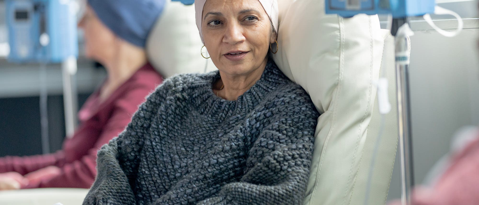 Zwei Frauen, die vordere mit einer hellen Mütze, die hintere mit einer blauen Mütze, sitzen auf Behandlungsstühlen in einer onkologischen Klinik.