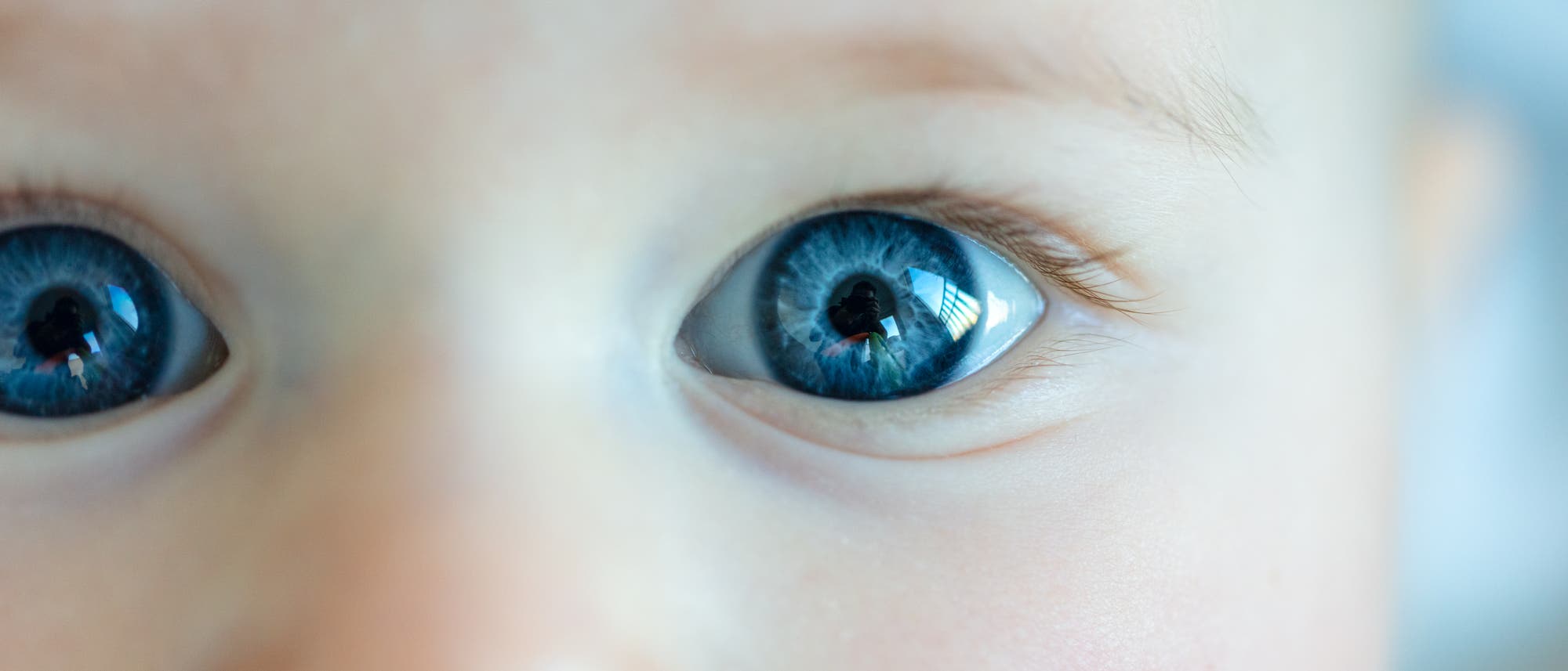 Nahaufnahme eines Babys mit großen, leuchtend blauen Augen, die direkt in die Kamera blicken. Das Bild zeigt die Details der Iris und die Reflexionen im Auge. Der Hintergrund ist unscharf, wodurch der Fokus auf dem Gesichtsausdruck des Babys liegt.
