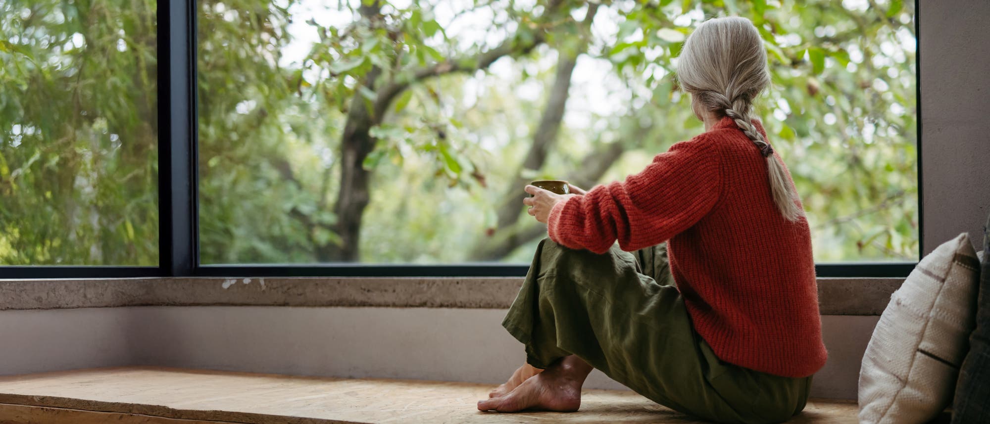Eine Person sitzt auf einer Holzbank vor einem gro&szlig;en Fenster und h&auml;lt eine Tasse. Sie tr&auml;gt einen roten Pullover und gr&uuml;ne Hosen. Drau&szlig;en sind gr&uuml;ne B&auml;ume zu sehen, die eine ruhige und entspannte Atmosph&auml;re schaffen. Ein Kissen liegt neben der Person.