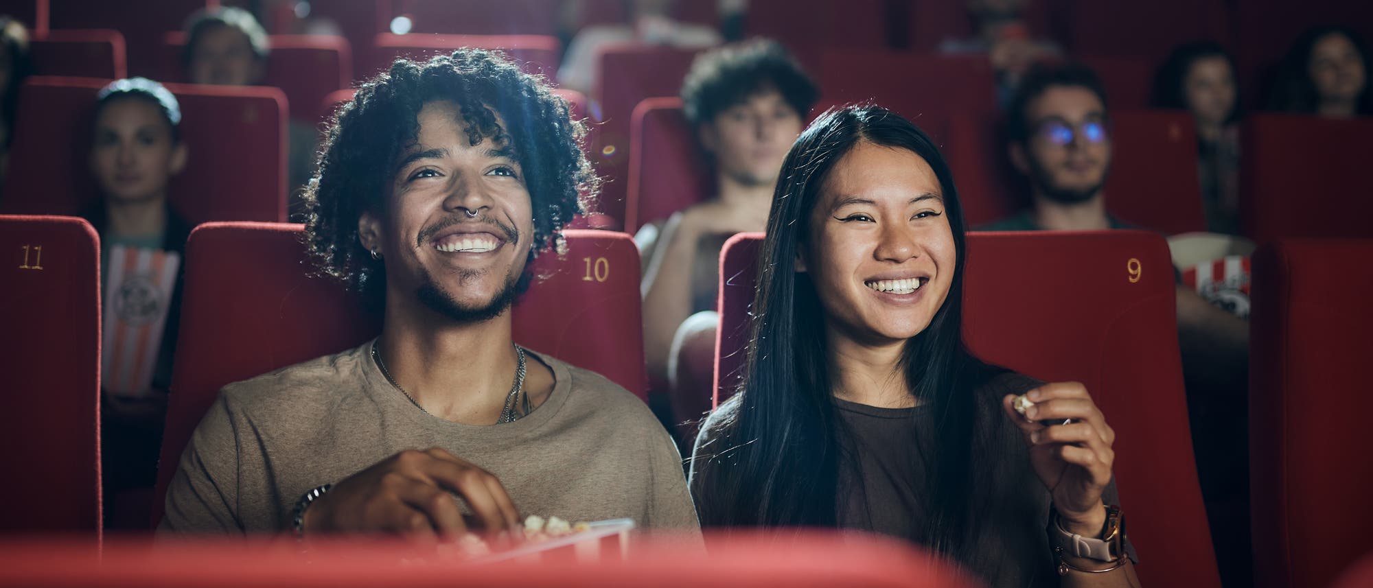 Eine Gruppe von Menschen sitzt in einem Kino auf roten Sitzen, die mit Nummern versehen sind. Zwei Personen im Vordergrund lächeln und halten Popcorn, während sie den Film genießen. Die Atmosphäre ist entspannt und fröhlich, mit gedämpftem Licht im Hintergrund.