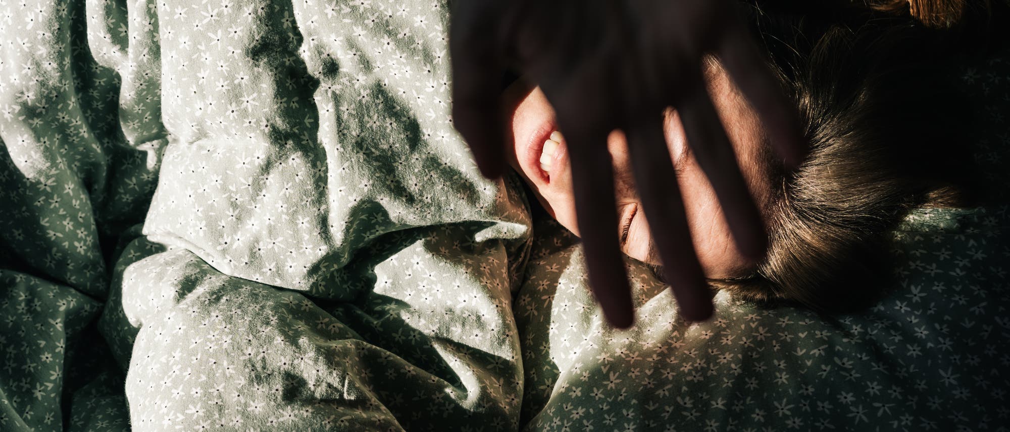 Eine Person liegt in einem Bett mit gemusterter Bettwäsche und hält eine Hand schützend vor das Gesicht, während Sonnenlicht auf sie fällt. Die Szene vermittelt ein Gefühl von Morgenruhe und Geborgenheit.