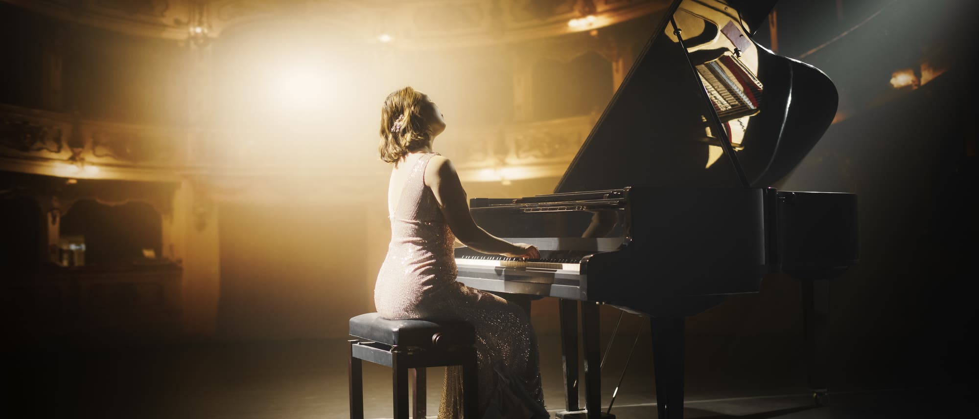 Eine Person in einem eleganten Kleid spielt Klavier auf einer B&uuml;hne in einem stimmungsvoll beleuchteten Theater. Der Fl&uuml;gel ist schwarz und gl&auml;nzend, und das Licht f&auml;llt sanft auf die Szene, wodurch eine warme, einladende Atmosph&auml;re entsteht. Im Hintergrund sind die R&auml;nge des Theaters zu sehen, die in ged&auml;mpftem Licht gehalten sind.