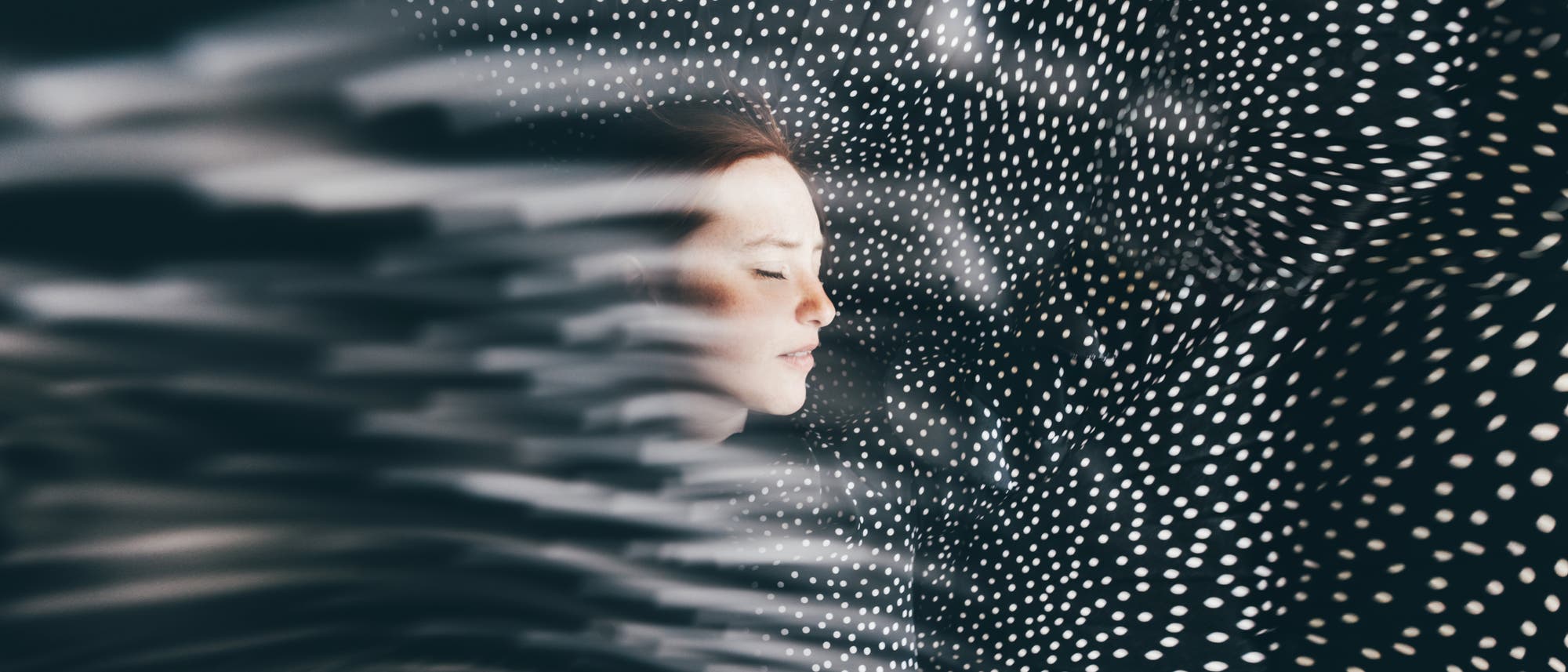 Eine Person mit geschlossenen Augen liegt in einem Bett, umgeben von einem dunklen Stoff mit wei&szlig;en Punkten. Der Stoff erzeugt einen wellenartigen Effekt, der das Bild dynamisch erscheinen l&auml;sst. Die Szene vermittelt Ruhe und Geborgenheit.