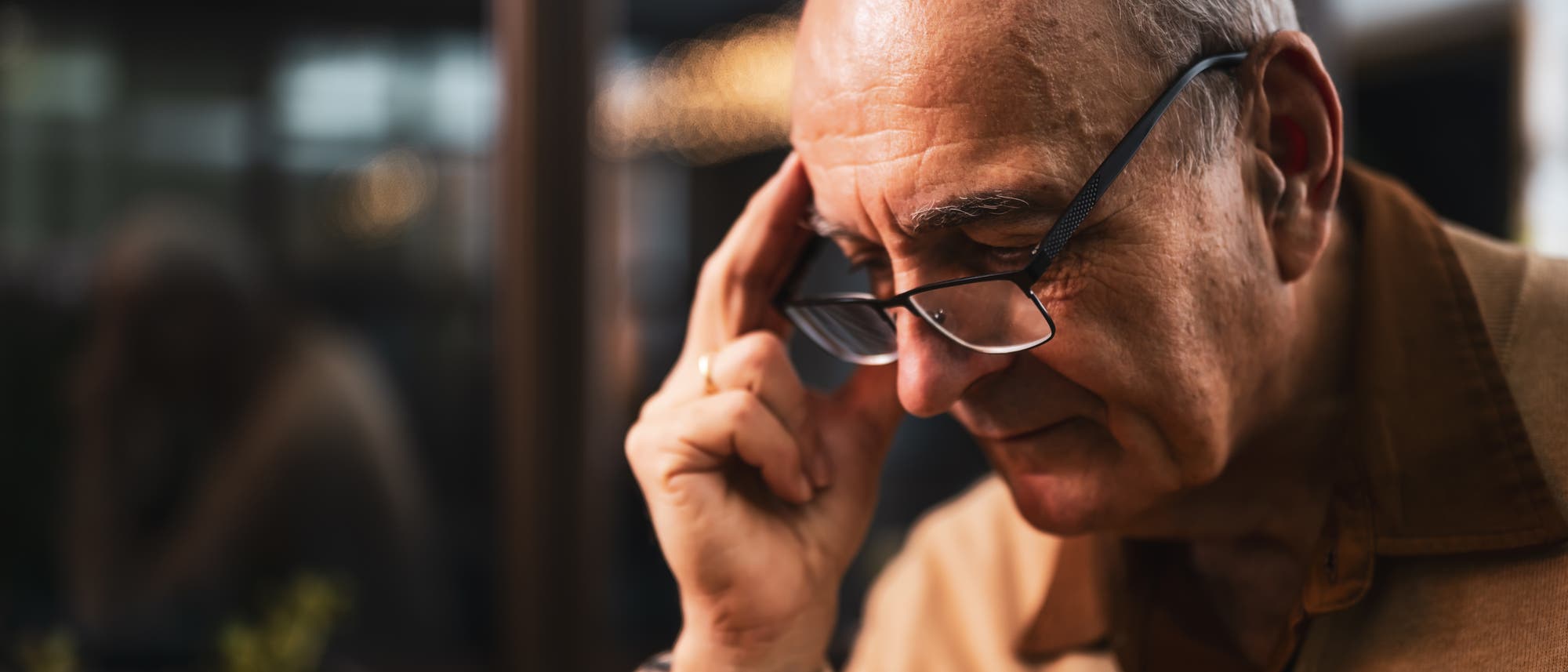 Ein älterer Mann mit Brille sitzt nachdenklich in einem Innenraum. Er trägt einen beigen Pullover und hält seine Brille mit der rechten Hand. Im Hintergrund sind verschwommene Fenster zu sehen. Ein älterer Mann mit Brille sitzt nachdenklich in einem Innenraum. Er trägt einen beigen Pullover und hält seine Brille mit der rechten Hand. Im Hintergrund sind verschwommene Fenster zu sehen.