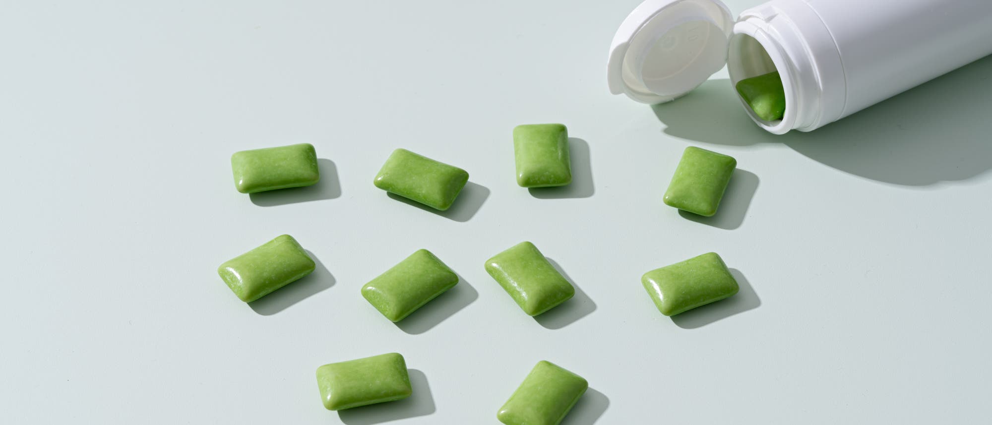 Eine weiße Plastikdose liegt auf einer hellen Oberfläche, aus der mehrere grüne Kaugummis herausgefallen sind. Die Kaugummis sind rechteckig und glänzend. Die Dose ist geöffnet und zeigt einen weiteren Kaugummi im Inneren. Die Szene ist minimalistisch und sauber gestaltet.
