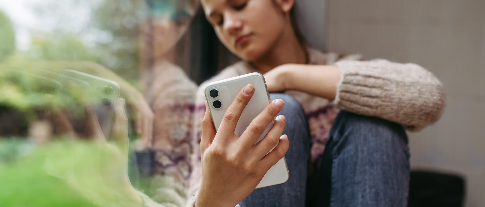 Eine Person sitzt nachdenklich an einem Fenster und h&auml;lt ein Smartphone in der Hand. Ihr Blick ist auf das Ger&auml;t gerichtet, w&auml;hrend ihr Spiegelbild im Glas sichtbar ist. Sie tr&auml;gt einen Strickpullover und Jeans. Drau&szlig;en ist unscharf eine gr&uuml;ne Landschaft zu erkennen.