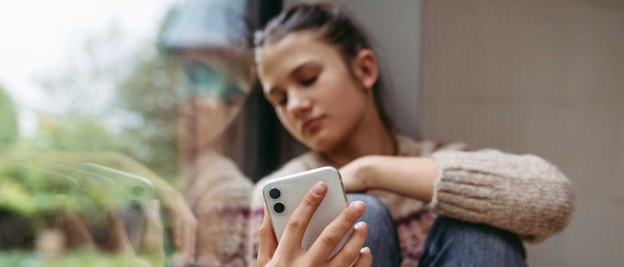 Eine Person sitzt nachdenklich an einem Fenster und hält ein Smartphone in der Hand. Die Reflexion der Person ist im Glas sichtbar. Die Szene vermittelt einen ruhigen, nachdenklichen Moment. Eine Person sitzt nachdenklich an einem Fenster und hält ein Smartphone in der Hand. Die Reflexion der Person ist im Glas sichtbar. Die Szene vermittelt einen ruhigen, nachdenklichen Moment.