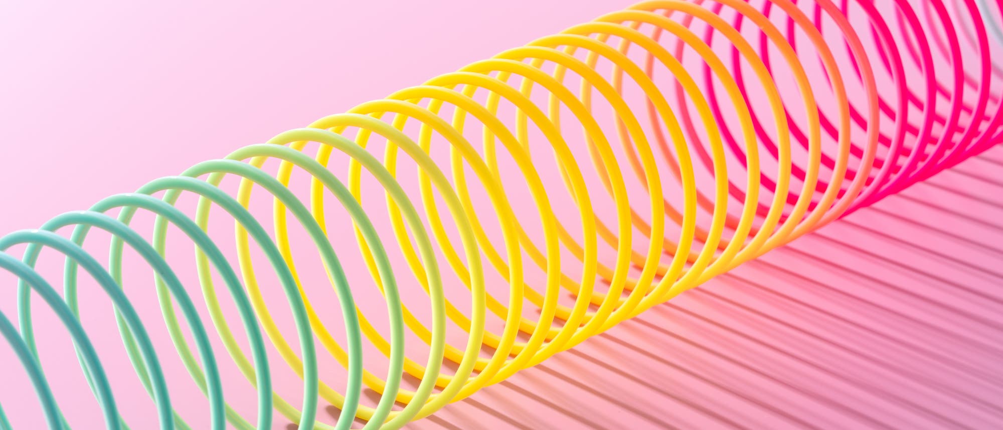 Ein farbenfrohes Spielzeug, bekannt als Slinky, liegt diagonal auf einer rosa Oberfl&auml;che. Die Spirale wechselt von T&uuml;rkis &uuml;ber Gelb zu Pink und wirft einen Schatten, der die Farben auf der Oberfl&auml;che reflektiert. Der Hintergrund ist in einem sanften Rosa gehalten.