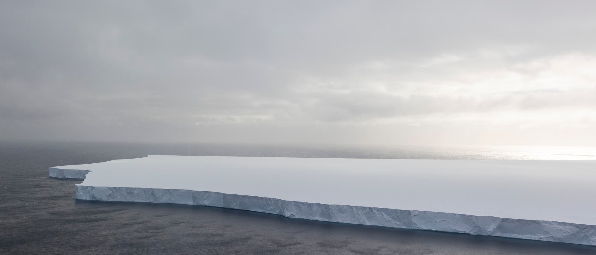 Ein großer, flacher Eisberg schwimmt im offenen Meer unter einem bewölkten Himmel. Die Oberfläche des Eisbergs ist glatt und weiß, während die Seiten steil ins Wasser abfallen. Das Meer ist dunkel und ruhig, und der Himmel zeigt diffuse Sonnenstrahlen, die durch die Wolken brechen. Ein großer, flacher Eisberg schwimmt im offenen Meer unter einem bewölkten Himmel. Die Oberfläche des Eisbergs ist glatt und weiß, während die Seiten steil ins Wasser abfallen. Das Meer ist dunkel und ruhig, und der Himmel zeigt diffuse Sonnenstrahlen, die durch die Wolken brechen.