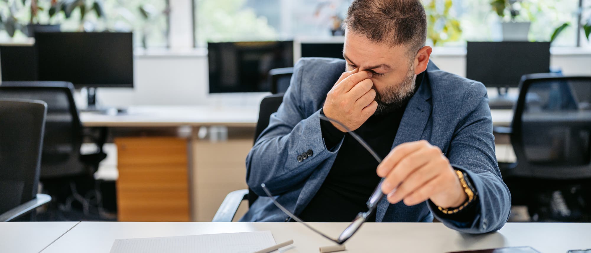 Ein Mann sitzt in einem Büro an einem Schreibtisch und reibt sich die Augen, während er eine Brille in der Hand hält. Auf dem Tisch liegen ein Notizbuch und ein Stift. Im Hintergrund sind Computer und Pflanzen zu sehen. Die Szene vermittelt den Eindruck von Müdigkeit oder Anstrengung bei der Arbeit.