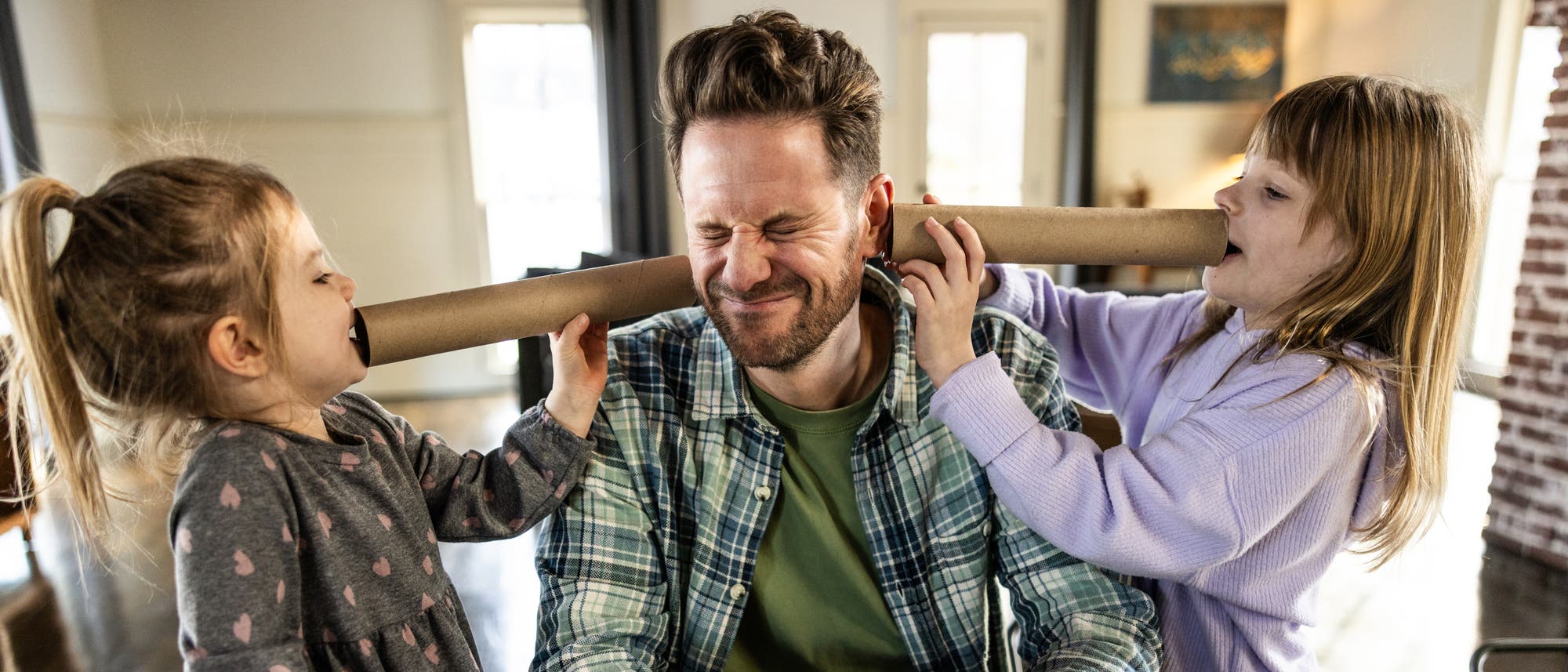 Ein Mann sitzt an einem Tisch. Zwei Kinder stehen neben ihm und halten ein Papprohr an seine Ohren, w&auml;hrend sie hineinsprechen. Der Mann kneift die Augen zusammen und l&auml;chelt leicht. Die Szene spielt in einem hellen Raum mit Fenstern im Hintergrund.
