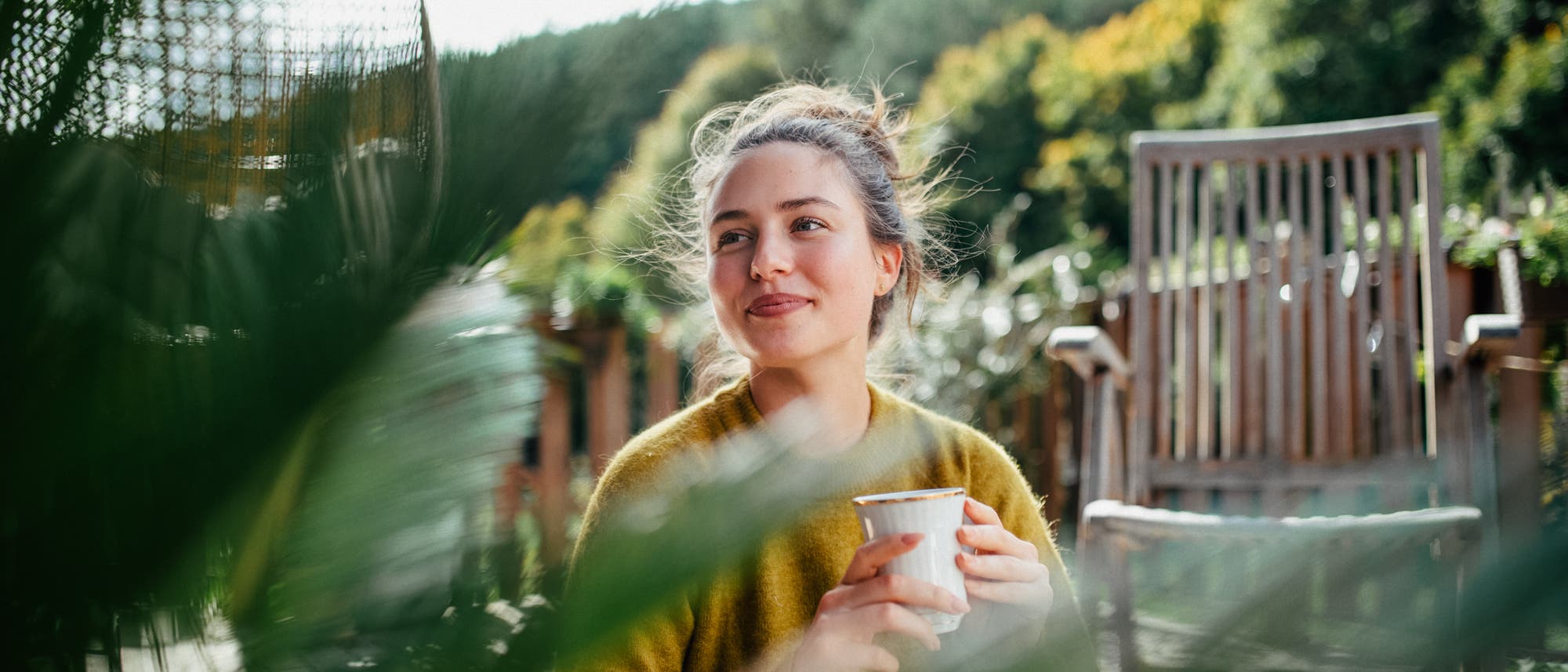 Eine Person sitzt entspannt auf einer Terrasse, umgeben von grünen Pflanzen. Sie hält eine Tasse in den Händen und lächelt leicht. Im Hintergrund sind ein Holzstuhl und eine üppige, bewaldete Landschaft zu sehen. Die Szene vermittelt eine ruhige und friedliche Atmosphäre. Eine Person sitzt entspannt auf einer Terrasse, umgeben von grünen Pflanzen. Sie hält eine Tasse in den Händen und lächelt leicht. Im Hintergrund sind ein Holzstuhl und eine üppige, bewaldete Landschaft zu sehen. Die Szene vermittelt eine ruhige und friedliche Atmosphäre.