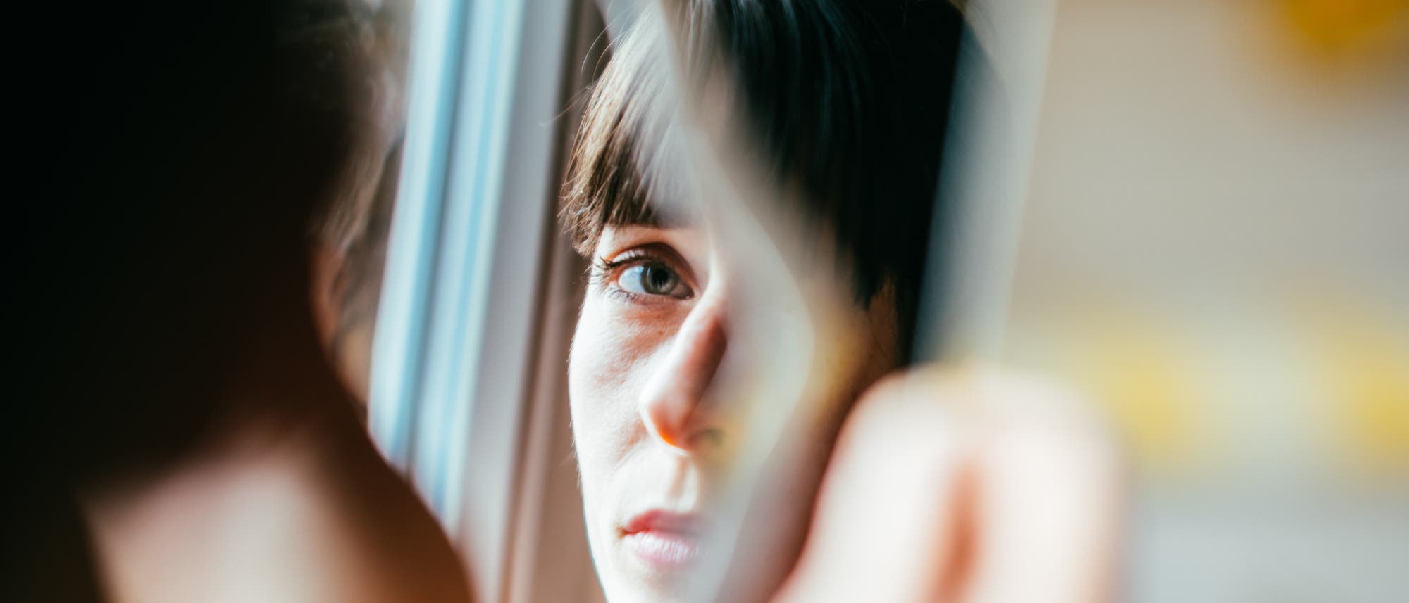 Eine Person schaut nachdenklich aus einem Fenster. Ihr Gesicht spiegelt sich im Glas wider, w&auml;hrend sie in die Ferne blickt. Die Szene vermittelt ein Gef&uuml;hl von Ruhe und Reflexion. Im Hintergrund sind verschwommene Details eines Innenraums zu erkennen.