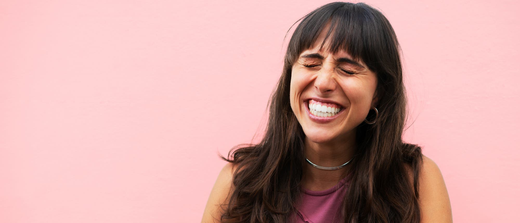 Eine Person mit langen Haaren lacht herzlich vor einem rosa Hintergrund. Sie tr&auml;gt ein &auml;rmelloses Oberteil und einen schlichten Halsreif. Die Augen sind geschlossen, und die Person wirkt fr&ouml;hlich und entspannt.