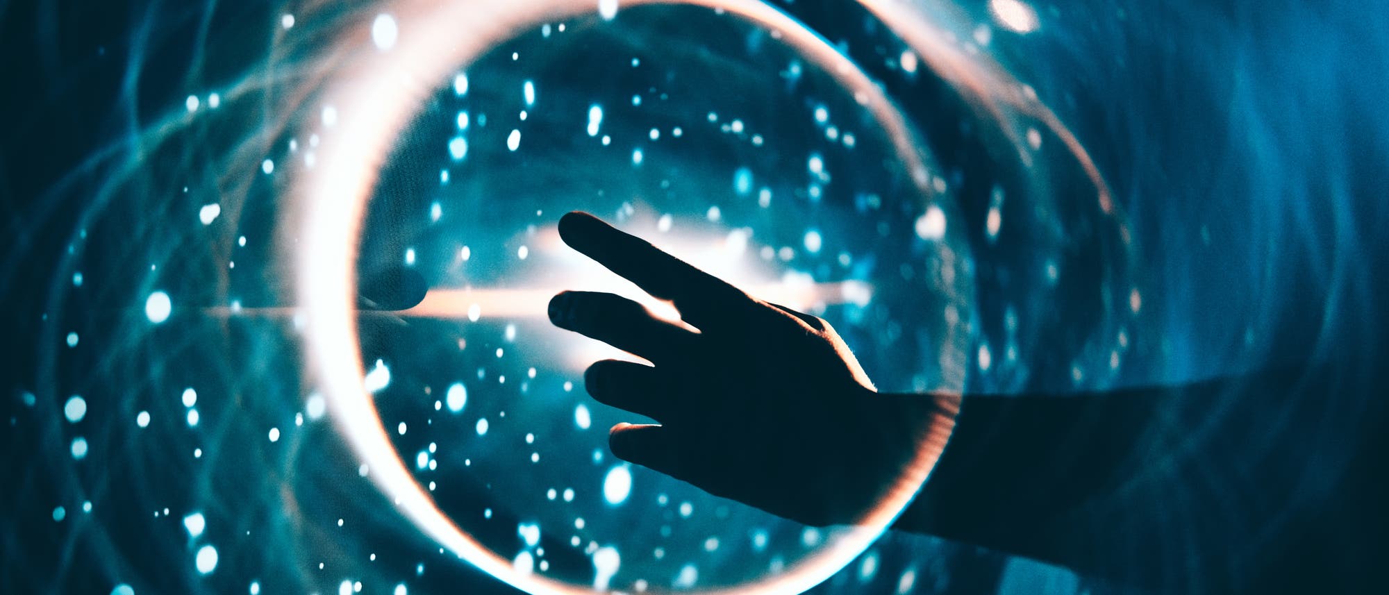 Eine Hand streckt sich in Richtung eines leuchtenden, kreisförmigen Lichts in einem dunklen, blauen Hintergrund. Um den Kreis herum sind helle, funkelnde Punkte verteilt, die an Sterne erinnern. Die Szene vermittelt ein Gefühl von Technologie und Magie. Eine Hand streckt sich in Richtung eines leuchtenden, kreisförmigen Lichts in einem dunklen, blauen Hintergrund. Um den Kreis herum sind helle, funkelnde Punkte verteilt, die an Sterne erinnern. Die Szene vermittelt ein Gefühl von Technologie und Magie.