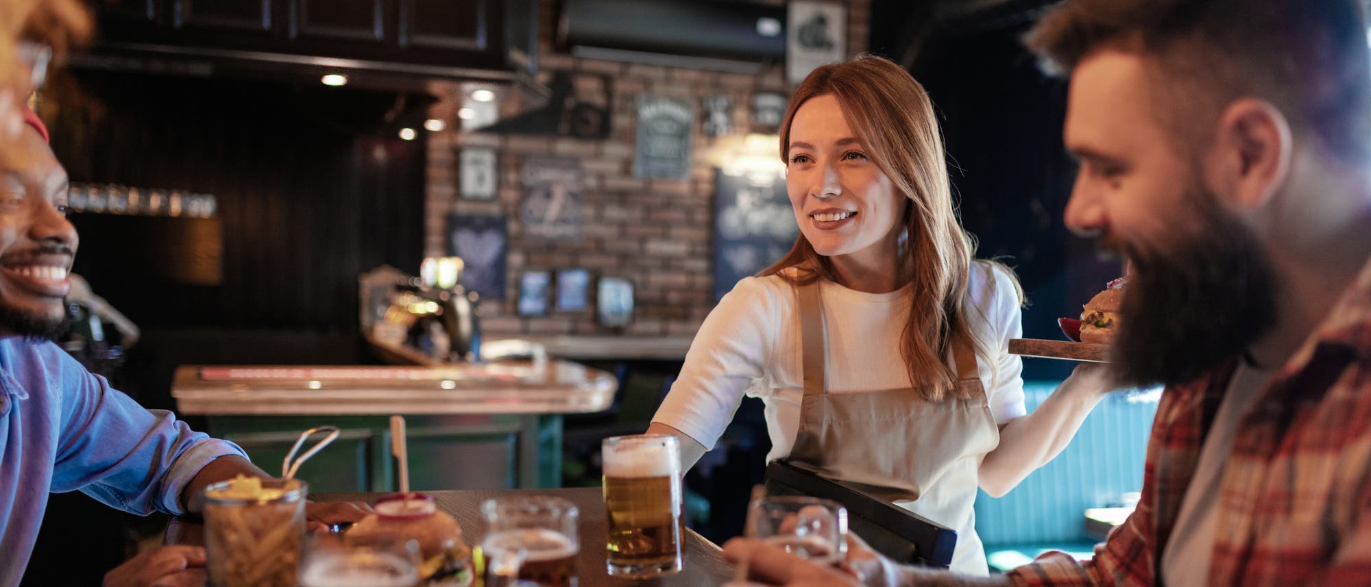 Eine Kellnerin in einem Café serviert einem Tisch mit drei Personen Getränke und Essen. Die Szene zeigt eine freundliche und einladende Atmosphäre in einem gemütlichen, rustikalen Ambiente mit Backsteinwänden. Auf dem Tisch stehen Gläser mit Getränken und ein Burger. Die Personen wirken entspannt und unterhalten sich.