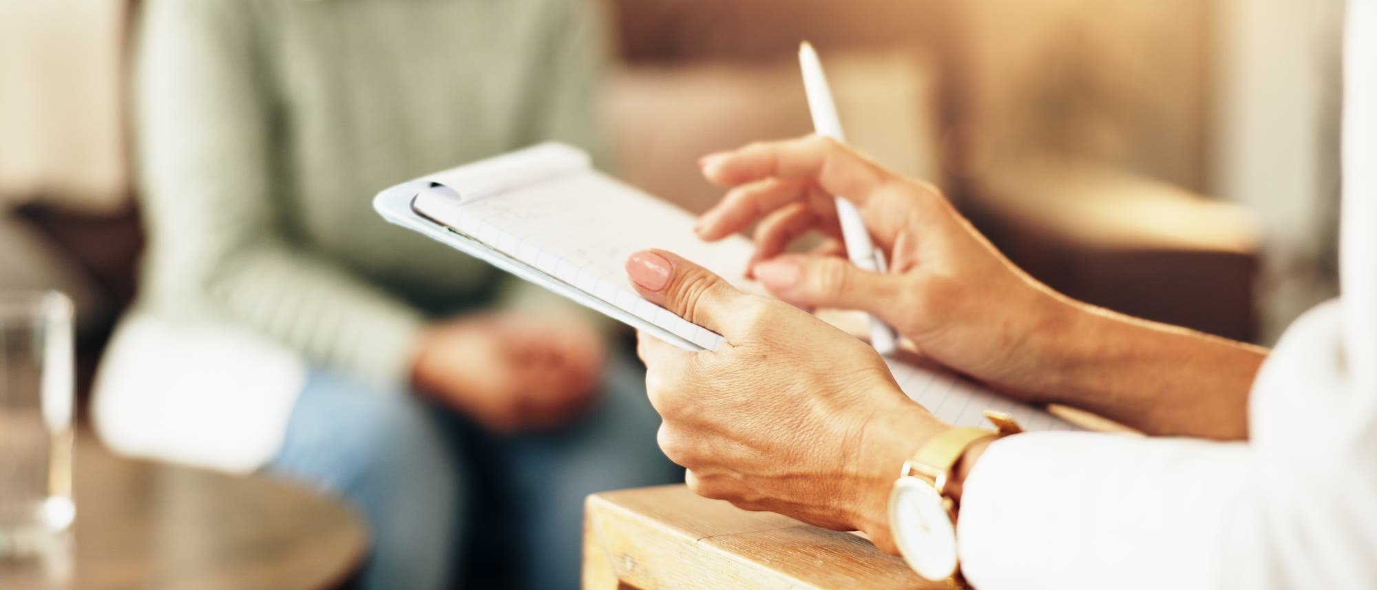 Eine Person h&auml;lt einen Notizblock und einen Stift in der Hand, w&auml;hrend sie mit einer anderen Person spricht, die im Hintergrund unscharf zu sehen ist. Die Szene wirkt wie ein Beratungsgespr&auml;ch oder ein Interview. Der Fokus liegt auf den H&auml;nden und dem Notizblock.