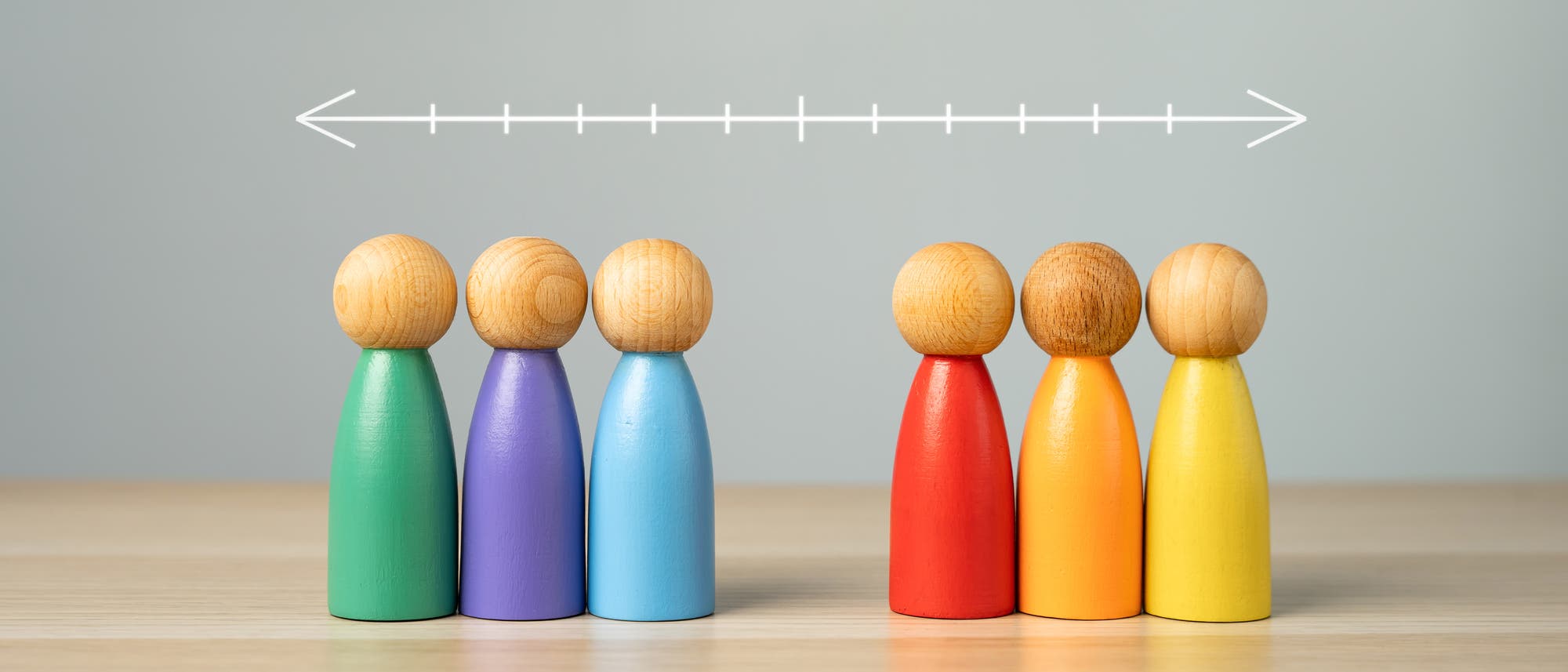 Sechs bunte Holzfiguren stehen auf einem Tisch, aufgeteilt in zwei Gruppen. Die linke Gruppe besteht aus drei Figuren in Grün, Lila und Blau, die rechte aus drei Figuren in Rot, Orange und Gelb. Über den Figuren ist ein horizontaler Pfeil mit Strichen, der auf die Trennung hinweist. Der Hintergrund ist grau. Sechs bunte Holzfiguren stehen auf einem Tisch, aufgeteilt in zwei Gruppen. Die linke Gruppe besteht aus drei Figuren in Grün, Lila und Blau, die rechte aus drei Figuren in Rot, Orange und Gelb. Über den Figuren ist ein horizontaler Pfeil mit Strichen, der auf die Trennung hinweist. Der Hintergrund ist grau.