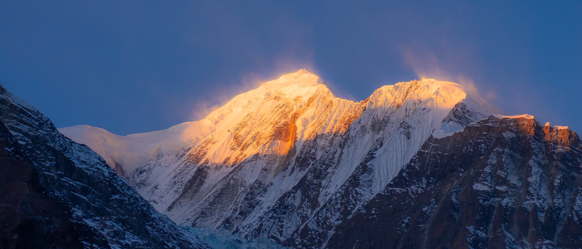 Ein schneebedeckter Berggipfel wird von der aufgehenden Sonne in warmes, goldenes Licht getaucht. Der Himmel ist klar und tiefblau, während die Sonnenstrahlen die schroffen Felswände und die Schneedecke des Berges hervorheben. Die Szene vermittelt eine ruhige und majestätische Atmosphäre.