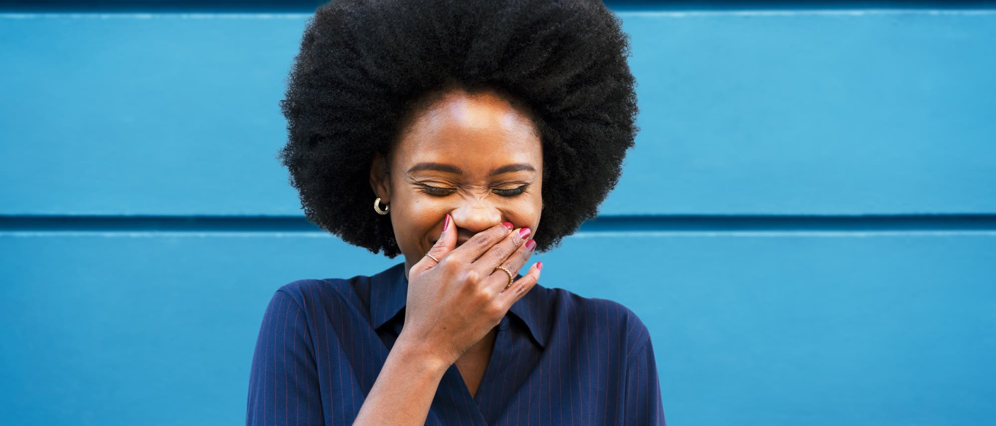 Eine Person steht vor einer blauen Wand und lacht, während sie sich die Hand vor den Mund hält. Sie trägt ein dunkelblaues Oberteil. Die Szene vermittelt Freude und Unbeschwertheit. Eine Person steht vor einer blauen Wand und lacht, während sie sich die Hand vor den Mund hält. Sie trägt ein dunkelblaues Oberteil. Die Szene vermittelt Freude und Unbeschwertheit.