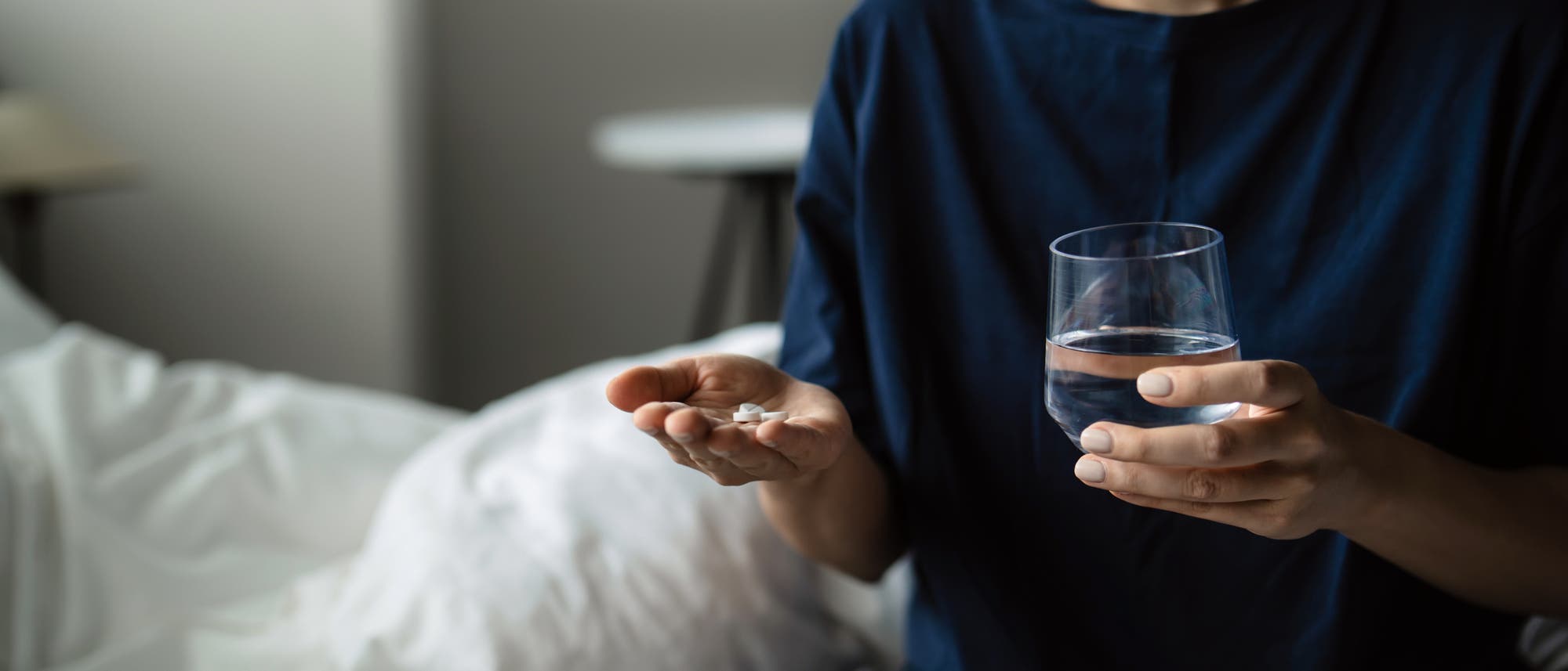 Eine Person sitzt auf einem Bett und h&auml;lt in der einen Hand ein Glas Wasser, in der anderen mehrere Tabletten. Sie tr&auml;gt ein dunkelblaues Oberteil und gestreifte Hosen. Im Hintergrund sind ein Kissen und ein Fenster zu sehen. Die Szene vermittelt den Eindruck, dass die Person sich darauf vorbereitet, die Tabletten einzunehmen.
