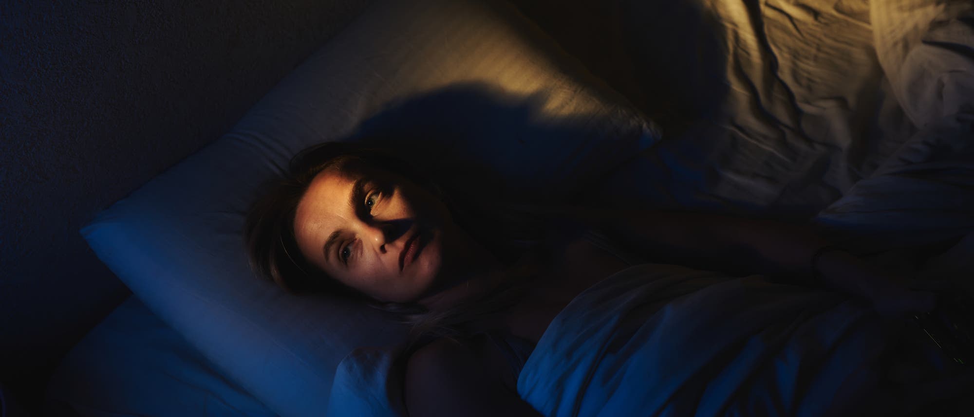 Eine Person liegt wach in einem Bett, umgeben von dunkler Beleuchtung. Ihr Gesicht ist teilweise von einem warmen Licht beleuchtet, während der Rest des Raumes in Schatten gehüllt ist. Die Bettwäsche ist weiß und der Ausdruck der Person wirkt nachdenklich.