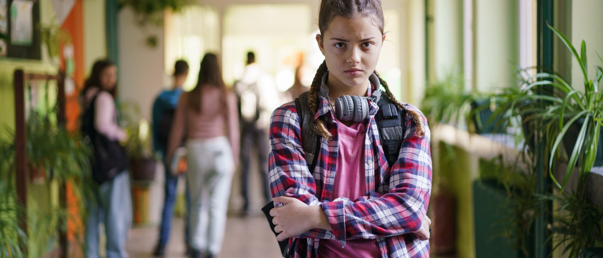 Ein junges Mädchen mit Kopfhörern um den Hals steht mit verschränkten Armen in einem Schulflur. Sie trägt ein kariertes Hemd und schaut ernst. Im Hintergrund sind unscharf andere Schüler zu sehen, die den Flur entlanggehen. Der Flur ist mit Pflanzen dekoriert und hat grüne Wände. Ein junges Mädchen mit Kopfhörern um den Hals steht mit verschränkten Armen in einem Schulflur. Sie trägt ein kariertes Hemd und schaut ernst. Im Hintergrund sind unscharf andere Schüler zu sehen, die den Flur entlanggehen. Der Flur ist mit Pflanzen dekoriert und hat grüne Wände.