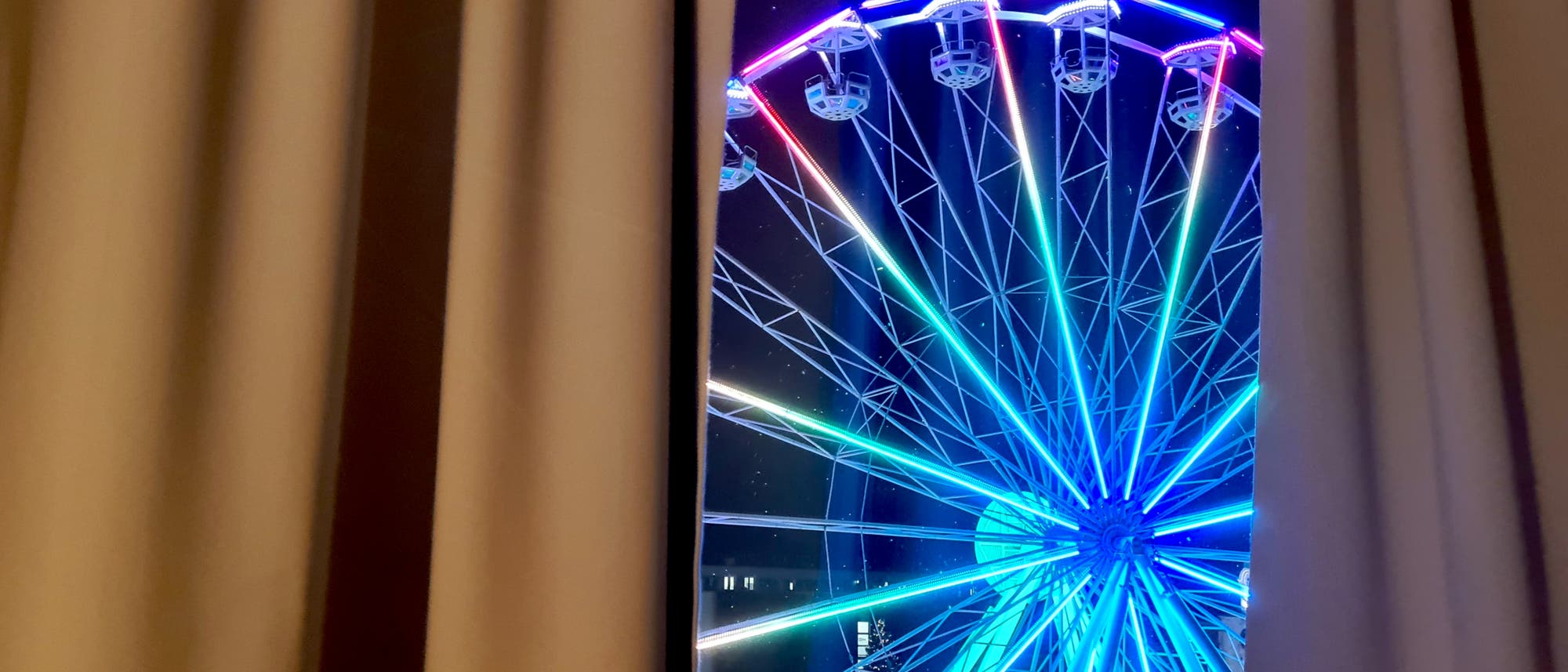 Ein beleuchtetes Riesenrad mit bunten Lichtern ist durch einen Spalt zwischen zwei Vorhängen sichtbar. Die Szene spielt sich nachts ab, und die Lichter des Riesenrads leuchten in verschiedenen Farben, darunter Blau, Grün und Rot. Im Hintergrund sind schwach beleuchtete Gebäude zu erkennen.