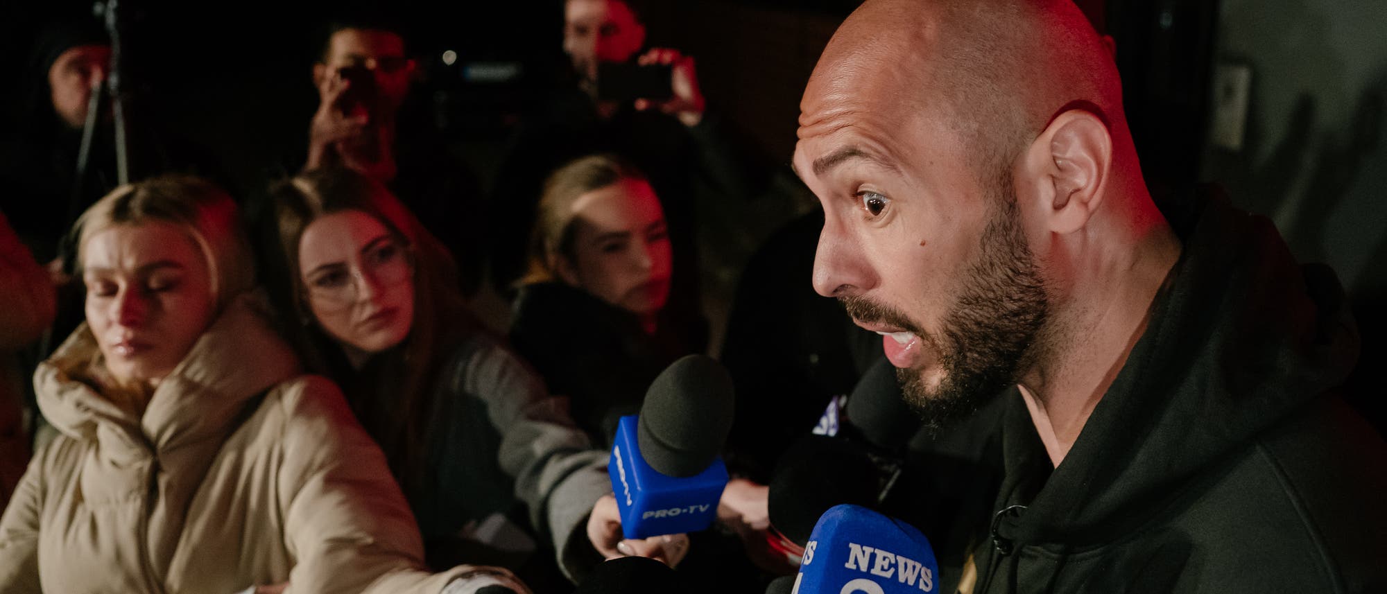 Eine Person mit Glatze und Vollbart spricht in mehrere Mikrofone von verschiedenen Nachrichtensendern, darunter "NEWS 24" und "PRO-TV". Um die Person herum stehen mehrere Reporter, die aufmerksam zuh&ouml;ren und die Szene mit Smartphones aufnehmen. Die Umgebung ist dunkel, was auf eine n&auml;chtliche Aufnahme hindeutet.