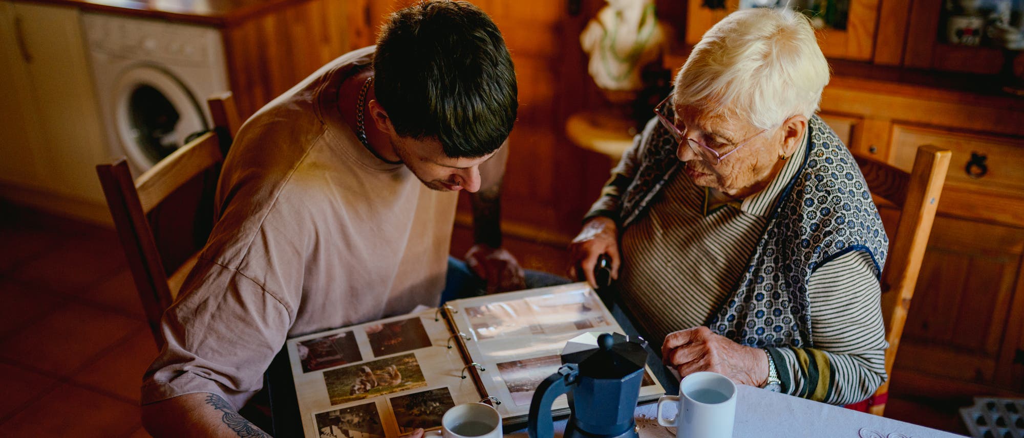 Eine ältere und eine jüngere Person sitzen an einem Tisch in einer gemütlichen Küche und betrachten gemeinsam ein Fotoalbum. Auf dem Tisch stehen zwei weiße Tassen und eine schwarze Kaffeekanne. Die Szene vermittelt eine warme, familiäre Atmosphäre. Eine ältere und eine jüngere Person sitzen an einem Tisch in einer gemütlichen Küche und betrachten gemeinsam ein Fotoalbum. Auf dem Tisch stehen zwei weiße Tassen und eine schwarze Kaffeekanne. Die Szene vermittelt eine warme, familiäre Atmosphäre.