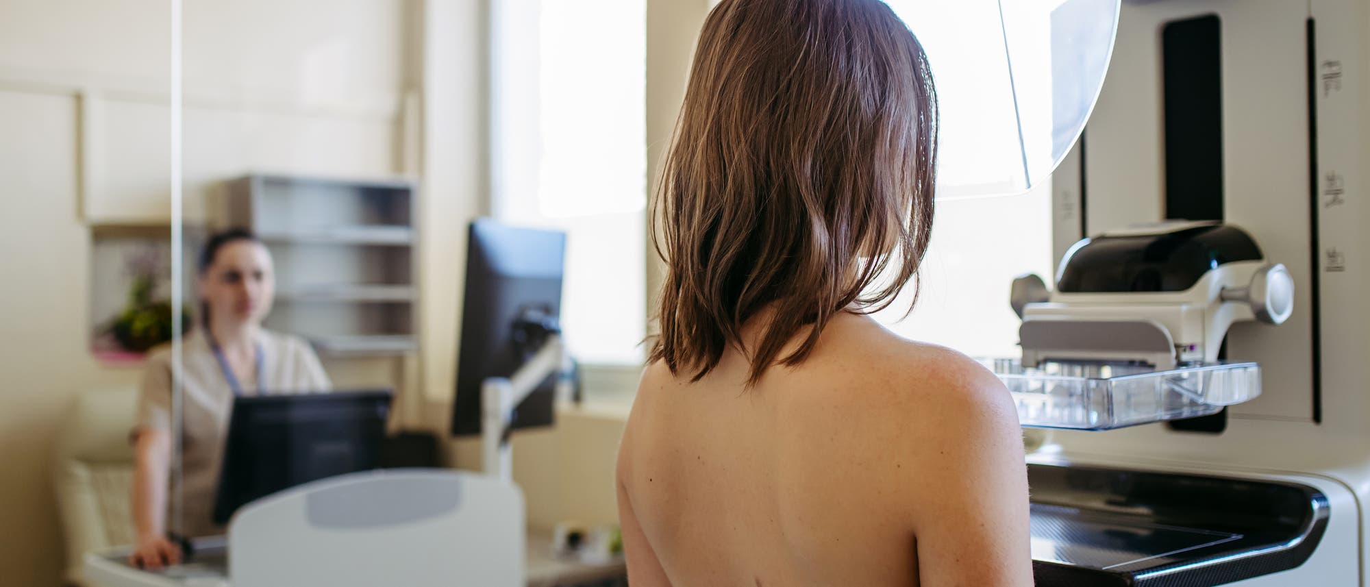 Eine Frau steht vor einem Mammographie-Gerät in einem medizinischen Untersuchungsraum. Im Hintergrund sitzt eine medizinische Fachkraft an einem Computer und beobachtet den Vorgang. Der Raum ist hell erleuchtet durch ein großes Fenster. Das Bild zeigt den Ablauf einer Brustkrebsfrüherkennung. Eine Frau steht vor einem Mammographie-Gerät in einem medizinischen Untersuchungsraum. Im Hintergrund sitzt eine medizinische Fachkraft an einem Computer und beobachtet den Vorgang. Der Raum ist hell erleuchtet durch ein großes Fenster. Das Bild zeigt den Ablauf einer Brustkrebsfrüherkennung.