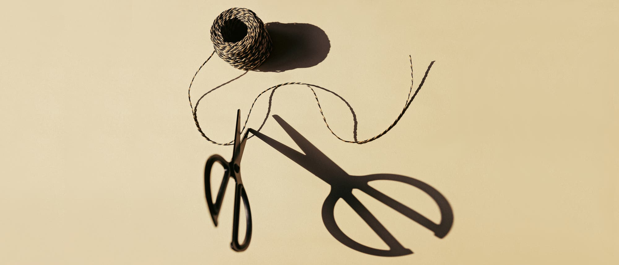 Eine Schere liegt auf einem beigen Hintergrund neben einer Rolle mit gedrehtem Garn. Der Schatten der Schere ist deutlich sichtbar und erstreckt sich über die Oberfläche. Die Schere ist kurz davor, das Garn durchzuschneiden. Das Garn ist teilweise abgewickelt und verläuft in einer geschwungenen Linie.
