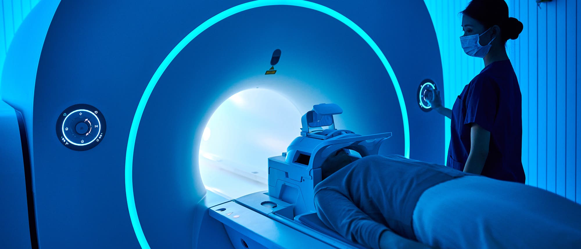 Eine Person liegt auf einer Liege, die in einen MRT-Scanner geschoben wird. Der Raum ist in blauem Licht beleuchtet. Eine medizinische Fachkraft steht daneben und &uuml;berwacht den Vorgang. Die Szene vermittelt eine klinische und professionelle Atmosph&auml;re.