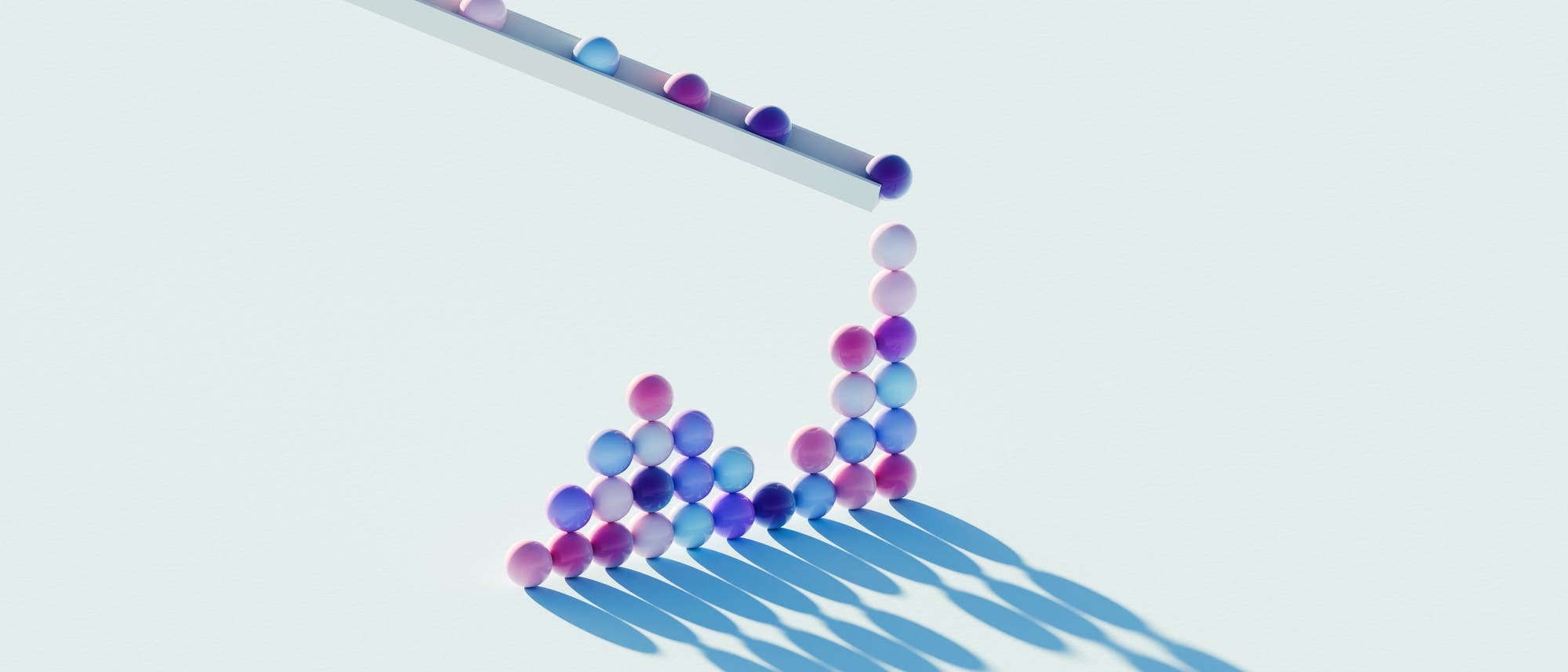 Eine 3D-Darstellung zeigt bunte Kugeln, die durch ein geneigtes Rohr rollen und sich am unteren Ende zu einer Pyramidenform stapeln. Die Kugeln sind in verschiedenen Pastellt&ouml;nen gehalten und werfen lange Schatten auf den hellen Hintergrund. Die Anordnung der Kugeln und ihre Schatten erzeugen ein geometrisches Muster.