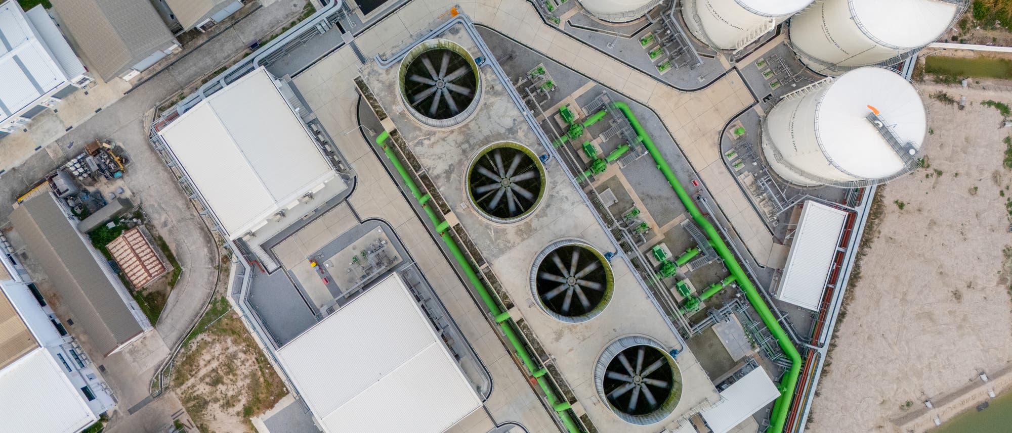 Luftaufnahme einer industriellen Anlage mit mehreren gro&szlig;en, runden Silos und rechteckigen Geb&auml;uden. In der Mitte befinden sich vier gro&szlig;e Ventilatoren, die Teil eines K&uuml;hlsystems zu sein scheinen. Die Anlage ist von Stra&szlig;en und Vegetation umgeben. Die Struktur und Anordnung der Geb&auml;ude und Silos deuten auf eine komplexe industrielle Nutzung hin.