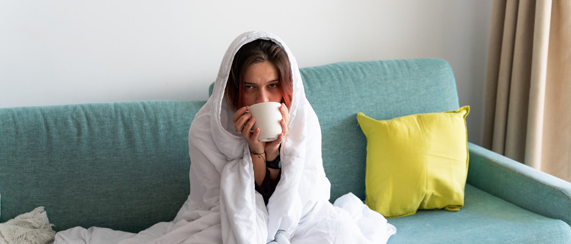 Eine Person sitzt auf einem t&uuml;rkisfarbenen Sofa, in eine wei&szlig;e Decke geh&uuml;llt, und h&auml;lt eine Tasse. Neben ihr liegt ein gelbes Kissen. Im Vordergrund ist ein Tisch mit einer Pflanze zu sehen. Die Szene vermittelt ein Gef&uuml;hl von Gem&uuml;tlichkeit und Entspannung.