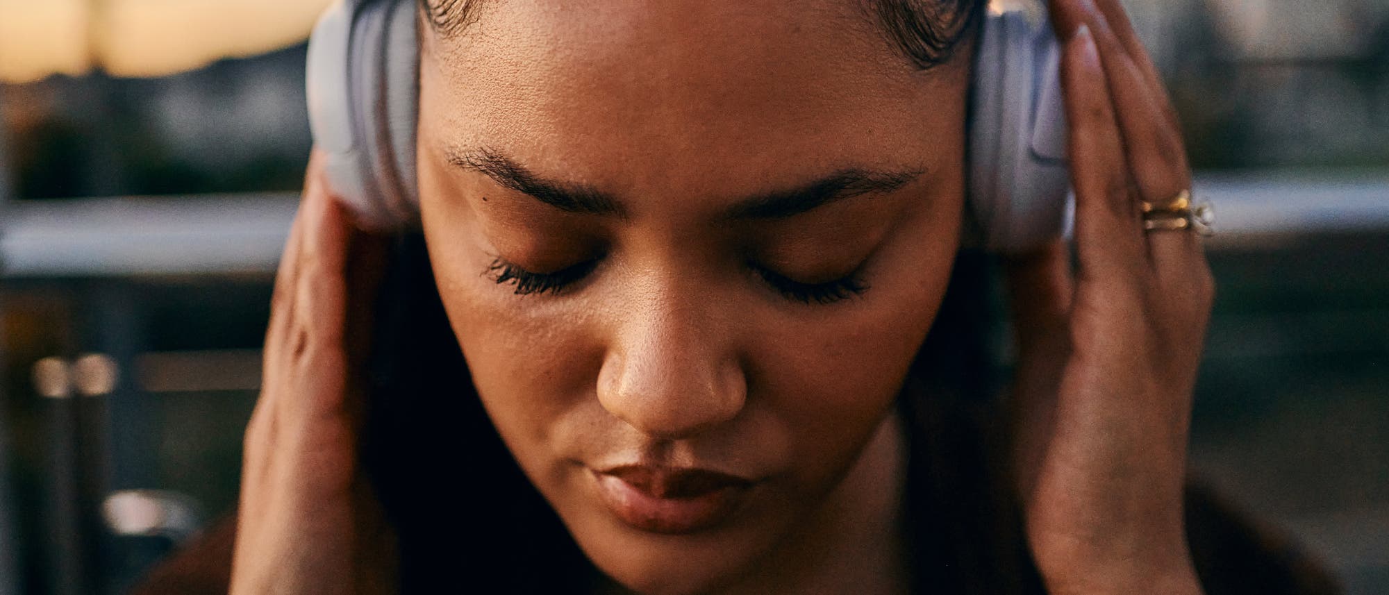 Eine Person tr&auml;gt Kopfh&ouml;rer und h&auml;lt sie mit beiden H&auml;nden fest. Die Augen sind geschlossen, was auf ein konzentriertes H&ouml;ren oder Genie&szlig;en von Musik hindeutet. Der Hintergrund ist unscharf, was den Fokus auf die Person lenkt. Die Szene findet bei Sonnenuntergang statt, was durch das warme Licht angedeutet wird.