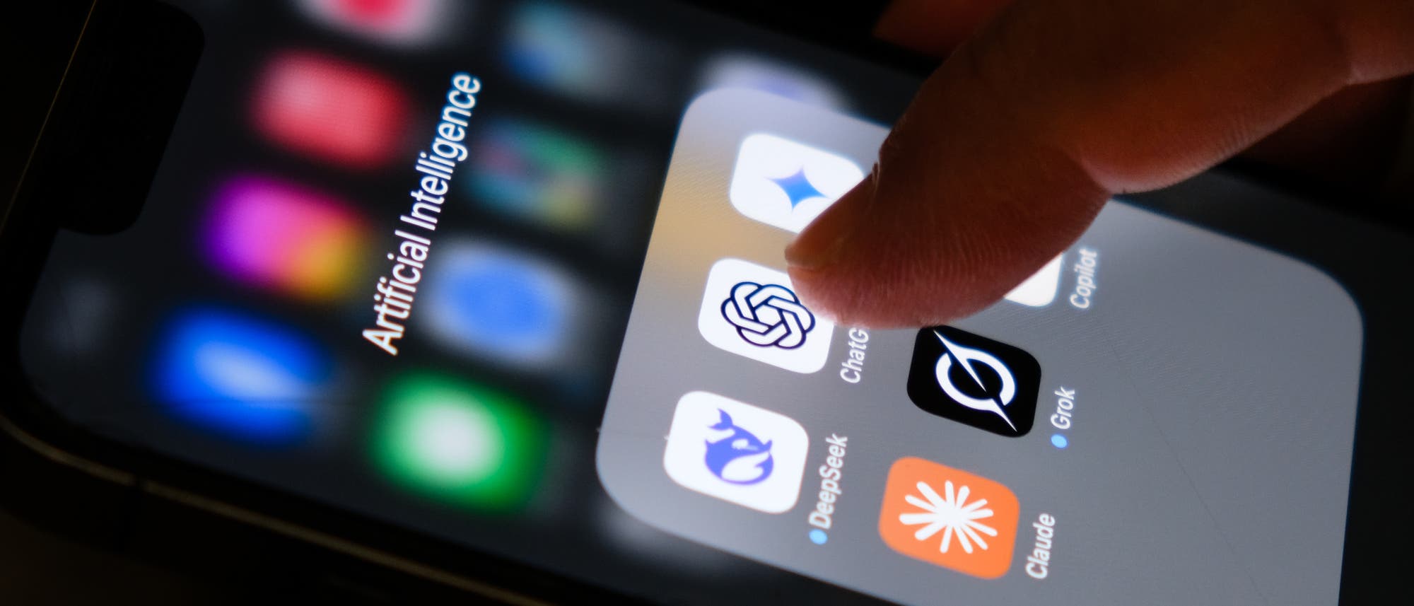 Ein Smartphone-Bildschirm zeigt einen Ordner mit dem Titel "Artificial Intelligence", der mehrere App-Icons enthält. Eine Hand berührt das Icon einer App mit einem spiralförmigen Logo. Die Apps im Ordner sind auf künstliche Intelligenz fokussiert. Der Hintergrund ist unscharf, um den Fokus auf die Interaktion mit der App zu lenken. Ein Smartphone-Bildschirm zeigt einen Ordner mit dem Titel "Artificial Intelligence", der mehrere App-Icons enthält. Eine Hand berührt das Icon einer App mit einem spiralförmigen Logo. Die Apps im Ordner sind auf künstliche Intelligenz fokussiert. Der Hintergrund ist unscharf, um den Fokus auf die Interaktion mit der App zu lenken.