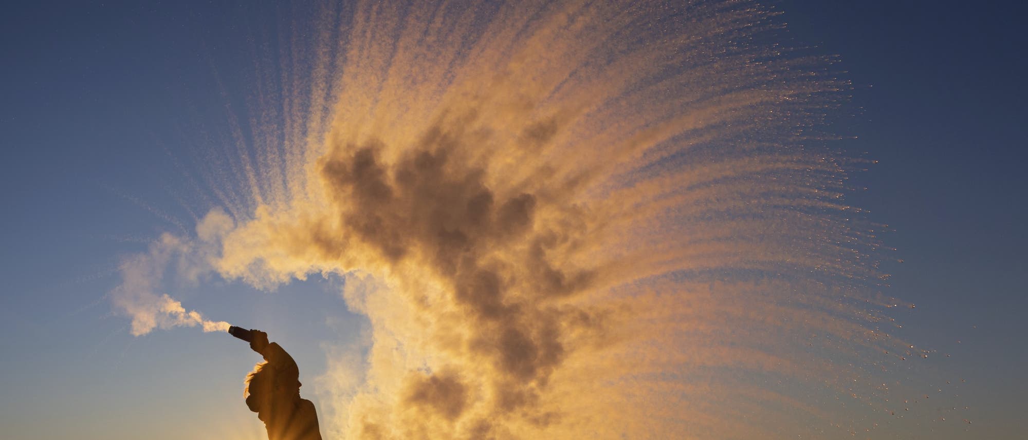 Eine Person steht auf einer schneebedeckten Fl&auml;che und wirft Wasser in die Luft, das in der kalten Luft zu einem beeindruckenden Bogen aus gefrorenem Nebel gefriert. Die tiefstehende Sonne im Hintergrund erzeugt einen leuchtenden Effekt, der die Szene dramatisch beleuchtet. Der Himmel ist klar und blau, was die winterliche Atmosph&auml;re verst&auml;rkt.