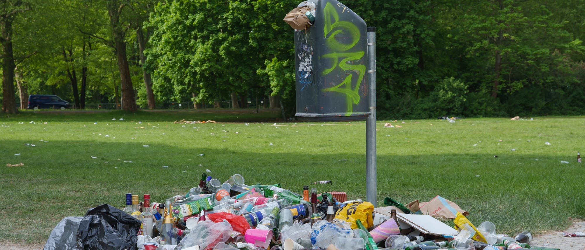 Ein &uuml;berf&uuml;llter M&uuml;lleimer in einem Park, umgeben von verstreutem M&uuml;ll wie Plastikflaschen, Pappbechern und Verpackungen. Im Hintergrund sind gr&uuml;ne B&auml;ume und eine Wiese zu sehen. Der M&uuml;lleimer ist mit Graffiti bespr&uuml;ht. Die Szene zeigt Umweltverschmutzung und die Notwendigkeit von mehr M&uuml;llentsorgung.