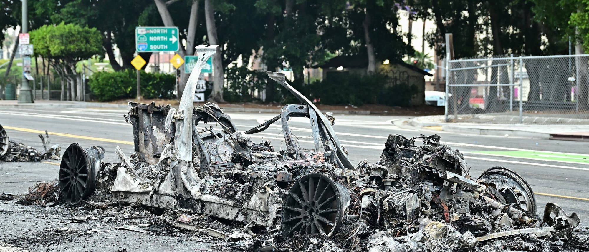 Ein ausgebranntes Autowrack steht auf einer Straße, umgeben von Trümmern. Im Hintergrund sind Straßenschilder zu sehen, die nach Norden und Süden weisen. Bäume und ein Zaun säumen die Straße. Ein ausgebranntes Autowrack steht auf einer Straße, umgeben von Trümmern. Im Hintergrund sind Straßenschilder zu sehen, die nach Norden und Süden weisen. Bäume und ein Zaun säumen die Straße.