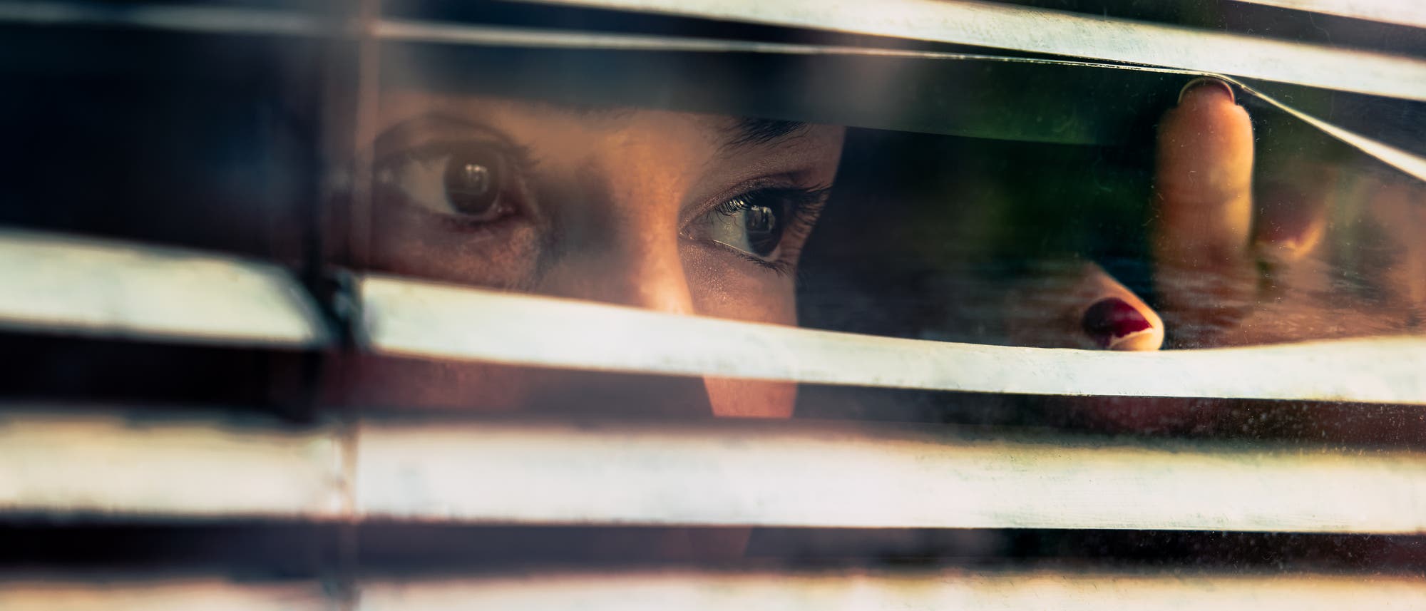 Eine Person schaut durch die Lamellen einer Jalousie. Nur die Augen und ein Teil des Gesichts sind sichtbar, während die Hand die Jalousie leicht anhebt. Die Szene vermittelt einen Eindruck von Neugier oder Vorsicht.