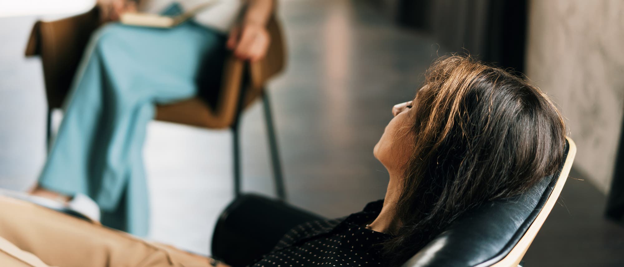 Eine Person liegt entspannt auf einer Couch in einem modernen Raum, w&auml;hrend eine andere Person in einem Stuhl sitzt und ein Notizbuch h&auml;lt. Die Szene deutet auf eine therapeutische Sitzung hin. Der Raum ist hell und minimalistisch eingerichtet.