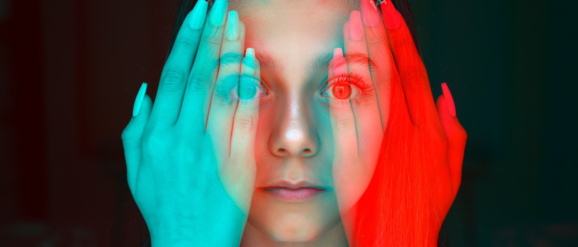 Eine Person h&auml;lt ihre H&auml;nde vor das Gesicht, wobei die Finger die Augen teilweise verdecken. Das Bild hat einen 3D-Effekt mit roten und blauen &Uuml;berlagerungen, die einen surrealen Eindruck erzeugen. Die Person blickt direkt in die Kamera.